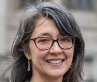 Sasha Welland headshot: shoulder-length grey cruly hair, red framed glasses, dangly earrings and smile showing teeth