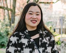 Full body shot of Christina Chung wearing long black and white coat, with hands crossed in front of her, smiling at the camera. Cherry blossom trees in the background. 