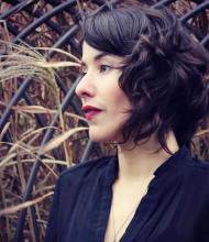 Left-facing profile of Michelle Morado wearing a black top, red lipstick, and with should-length curly brown hair. Background of lattice over hay. 