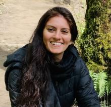 Upper body shot of a woman with long hair, wearing a black jacket with scarf, and smiling at the camera. Background is of a rock and tree with moss growing on it.