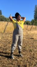 Keila Taylor wearing a yellow shirt and overalls standing in a field with her legs spread and firmly planted and her hands clasped behind her head. She's looking up at the sky. 