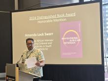 A person stands at a podium speaking, with a presentation slide behind them about the 2024 Distinguished Book Award Honorable Mention for "Envisioning African Intersex" by Amanda Lock Swarr.