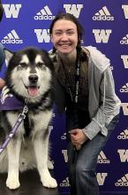 Hailey Capps crouched next to Dubs, an Alaskan Malamute, who is on a table to her right. They are in front of a purple backdrop covered in gold "W"s and the Adidas logo. 