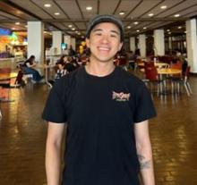 Upper body shot of Kai Mai in a black tshirt with a baseball cap on his head. Kai is a young Asian male with light skin and is smiling at the camera.