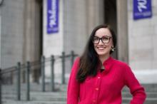 Upper body shot of Marika Cifor wearing red blouse and glasses with long brown hair over right-shoulder. UW's Suzzallo Hall is in the background and has purple "Welcome" banners draped on both sides of the entryway.