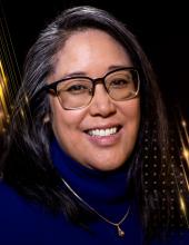 Headshot of Regina Yung Lee smiling. She has long black hair with grey streaks, square framed glasses, and is wearing a royal blue turtleneck sweater with a simple necklace on top. The background is black with gold rays.  