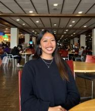 Thea Deanon, a young woman with olive-toned skin and long brown hair, sitting at a table in what appears to be a lunch room. She is wearing a dark sweatshirt, a thin necklace, and has her hands clasped in front of her. She is smiling at the camera. 