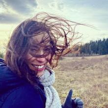 Upper body shop of woman smiling at camera, wearing winter jacket and scarf, with shoulder-length brown hair which is blowing in the wind and in her face. Background is beach and tree line. 