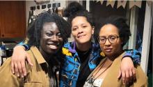 Upper body shot of three women of color (mostly) smiling at the camera. The woman in the middle has her arms around the other two. 