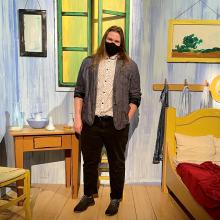 A man standing in the center of a stage designed to look like a bedroom with a chair, desk, bed, and artwork on the walls. The man with shoulder-length brown hair is wearing a sweater over a button-up shirt, black pants and shoes, and a mask on his face.