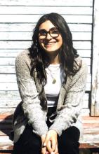 Kelen Tamurian, a young woman with long brown hair and glasses, sitting on a wooden porch with a lake in the background.