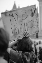 Gray scale image of the back of a person with shoulder-length hair holding a white sign with black handwriting that reads "My Body, My Choice"  