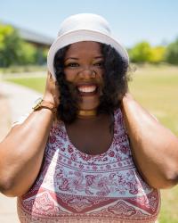 Keila Taylor smiling, wearing a white hat and patterned top, standing outdoors with green grass and trees in the background.