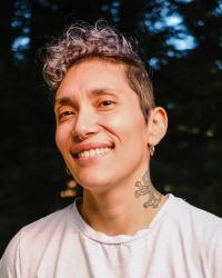 fabian romero standing in front of the woods wearing a pocket t-shirt, with tattoos on arms and neck, hair shaved on the sides and curly on top