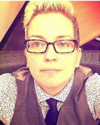 Jen Self sitting in a chair smiling up at the camera. They are wearing square-framed glasses and a vest with a tie over a printed dress shirt. 