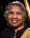 Headshot of Priti Ramamurthy smiling at the camera. Wearing glasses, gold and white beaded dangle earrings, and a black tunic with a flowered scarf on the right shoulder.  The background is black with gold stripes.