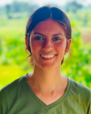 Nastasia is wearing an olive green t-shirt, standing against a light green background. 