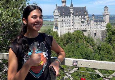 Upper body shot of Jordynn smiling at the camera and giving a thumbs up. She has long brown hair in a ponytail and is wearing a graphic t-shirt. In the background is Germany's famous Neuschwanstein Castle.