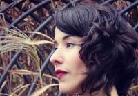 Left-facing profile of Michelle Morado wearing a black top, red lipstick, and with should-length curly brown hair. Background of lattice over hay. 