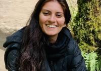 Upper body shot of a woman with long hair, wearing a black jacket with scarf, and smiling at the camera. Background is of a rock and tree with moss growing on it.