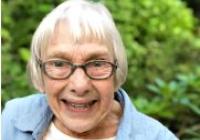 Photo of Carol Allen, an old white woman with ear-length white hair, wearing glasses, a blue button-up over a white t-shirt, with green foliage in the background