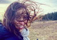 Upper body shop of woman smiling at camera, wearing winter jacket and scarf, with shoulder-length brown hair which is blowing in the wind and in her face. Background is beach and tree line. 