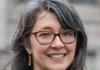 Sasha Welland headshot: shoulder-length grey cruly hair, red framed glasses, dangly earrings and smile showing teeth