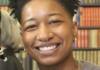 Headshot of Bobbi Kindred smiling at the camera. They have a short afro, and are wearing a thin gold chain and a grey sweater.