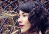 Left-facing profile of Michelle Morado wearing a black top, red lipstick, and with should-length curly brown hair. Background of lattice over hay. 