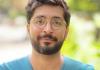 Upper body shot of Saad Khan smiling at the camera. He's wearing a light-blue v-neck t-shirt and glasses. The background is blurred greens indicating he may be outside.