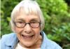 Photo of Carol Allen, an old white woman with ear-length white hair, wearing glasses, a blue button-up over a white t-shirt, with green foliage in the background