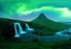 The Northern Lights glow in vibrant green over Kirkjufell Mountain in Iceland. A nearby waterfall cascades into a river, surrounded by lush greenery, with the iconic mountain peak rising in the background under the aurora-lit sky.