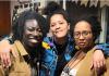 Upper body shot of three women of color (mostly) smiling at the camera. The woman in the middle has her arms around the other two. 