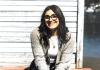 Kelen Tamurian, a young woman with long brown hair and glasses, sitting on a wooden porch with a lake in the background.