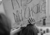 Gray scale image of the back of a person with shoulder-length hair holding a white sign with black handwriting that reads "My Body, My Choice"  