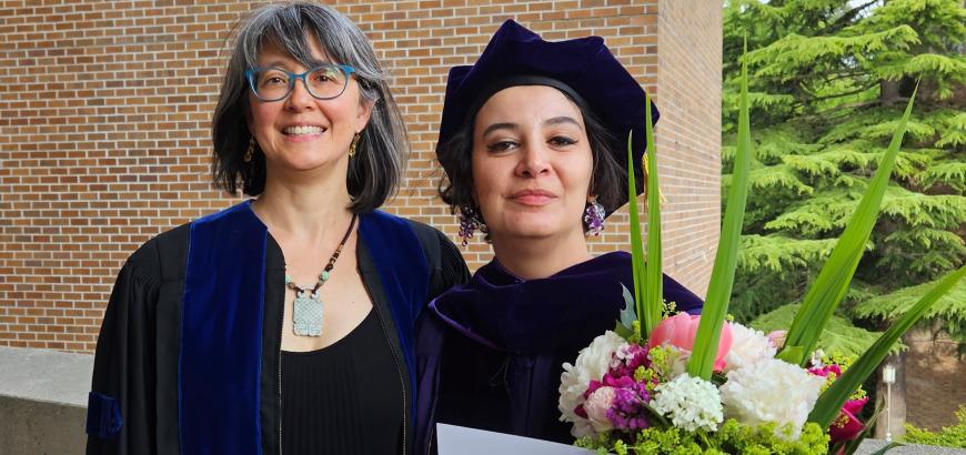 Two women, one in graduation gown and one in cap and gown, standing next to each other and smiling for the camera. The one in cap and gown, on the right, is holding a bouquet of flowers and a graduation certificate.