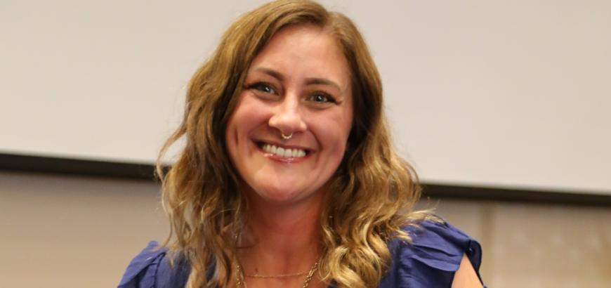 Upper body shot of a woman with long blond hair who is wearing a purple dress and smiling at the camera.