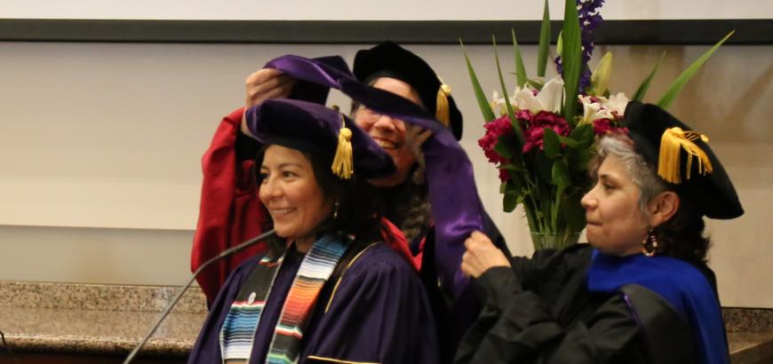 Profile image of three women in graduation caps and gowns. Two women are behind the third and are placing a graduation hood over her head.