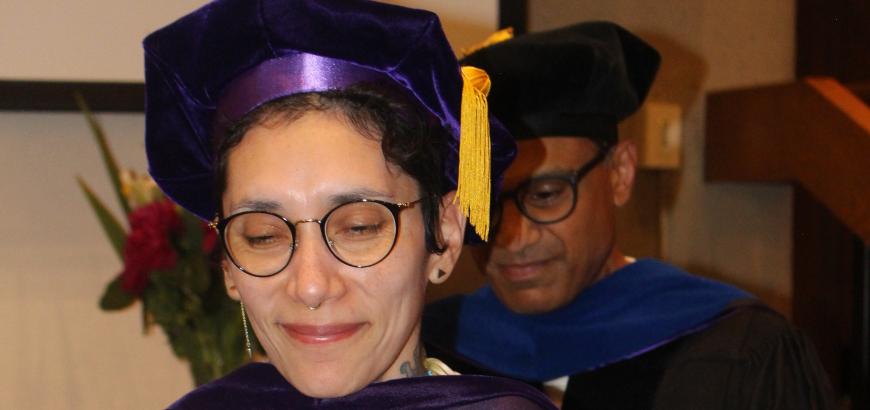 Upper body shot of two people in full graduation regalia. One is standing in front of the other as they receive their doctoral hood from the person behind them..